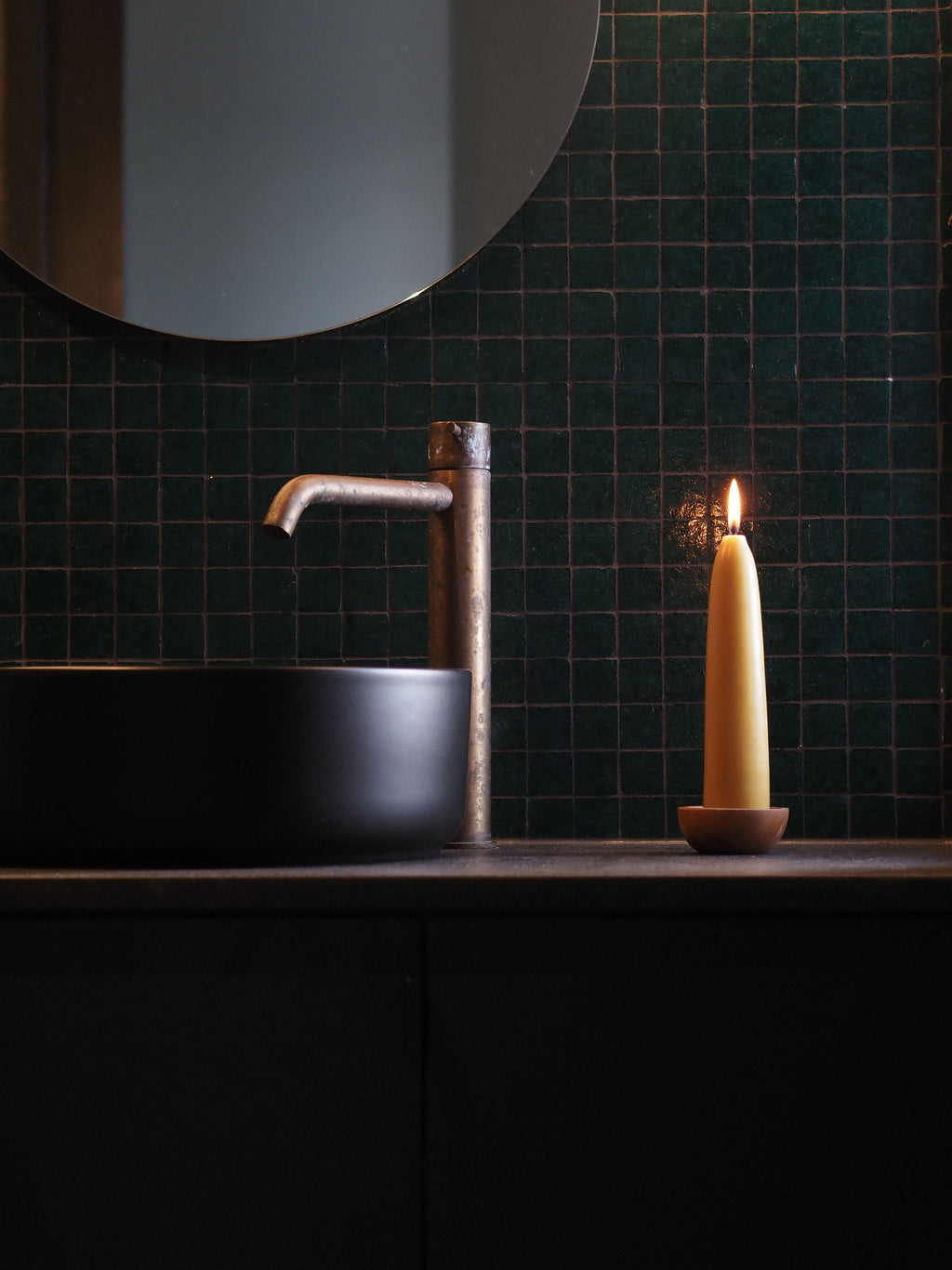 A modern bathroom scene with a matte black vessel sink, a tall brushed brass faucet, a lit beeswax candle on a wooden surface, and a dark green tiled wall background.