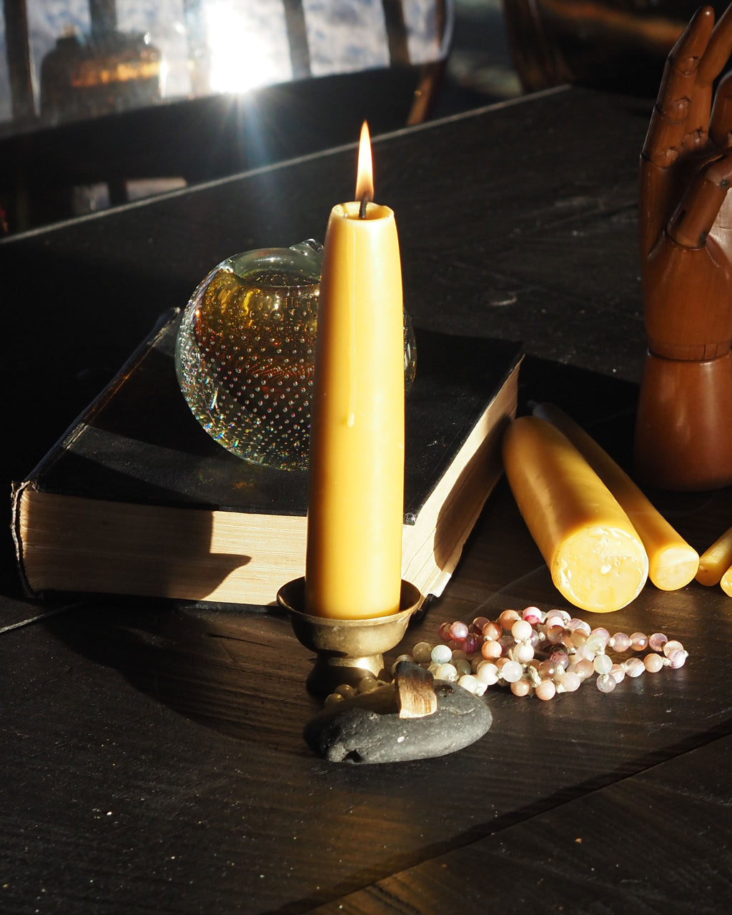 A lit beeswax taper candle stands on a dark surface, surrounded by a handmade glass ashtray, gemstone mala meditation beads, and additional beeswax candles.