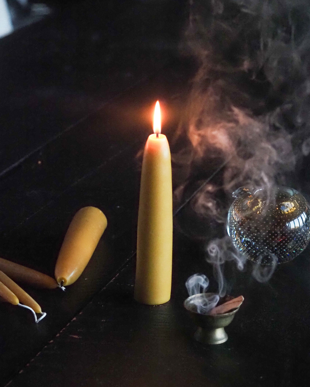 A lit beeswax taper candle stands on a dark surface, surrounded by smoky incense, a metallic incense holder, and additional beeswax candles.