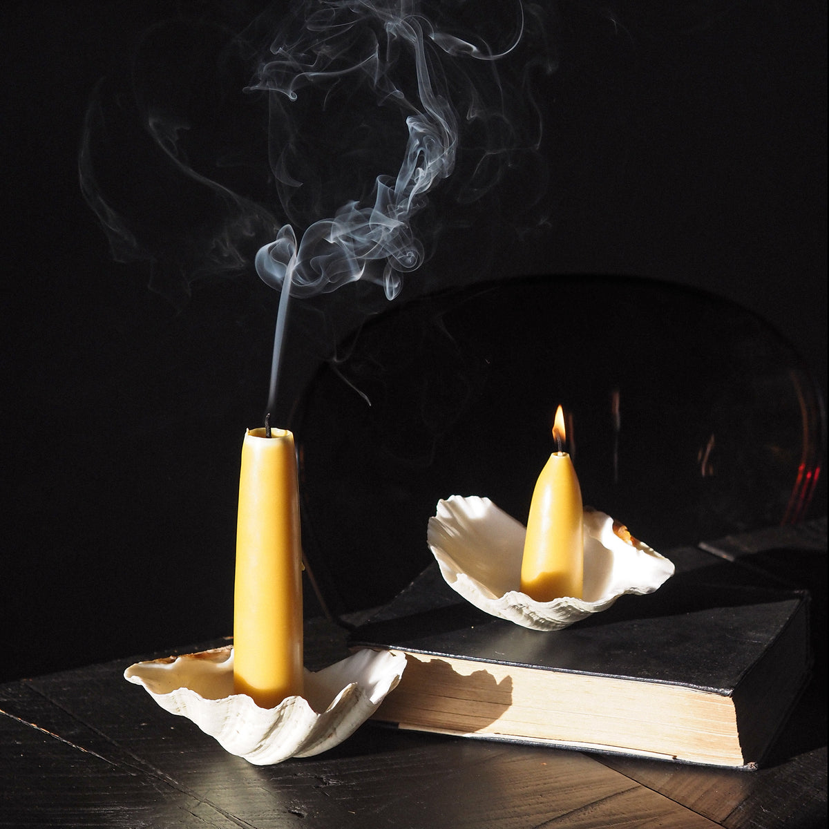 Two beeswax taper candles in white shell holders, one with smoke rising and one lit with a flame, arranged on a dark book against a black background.