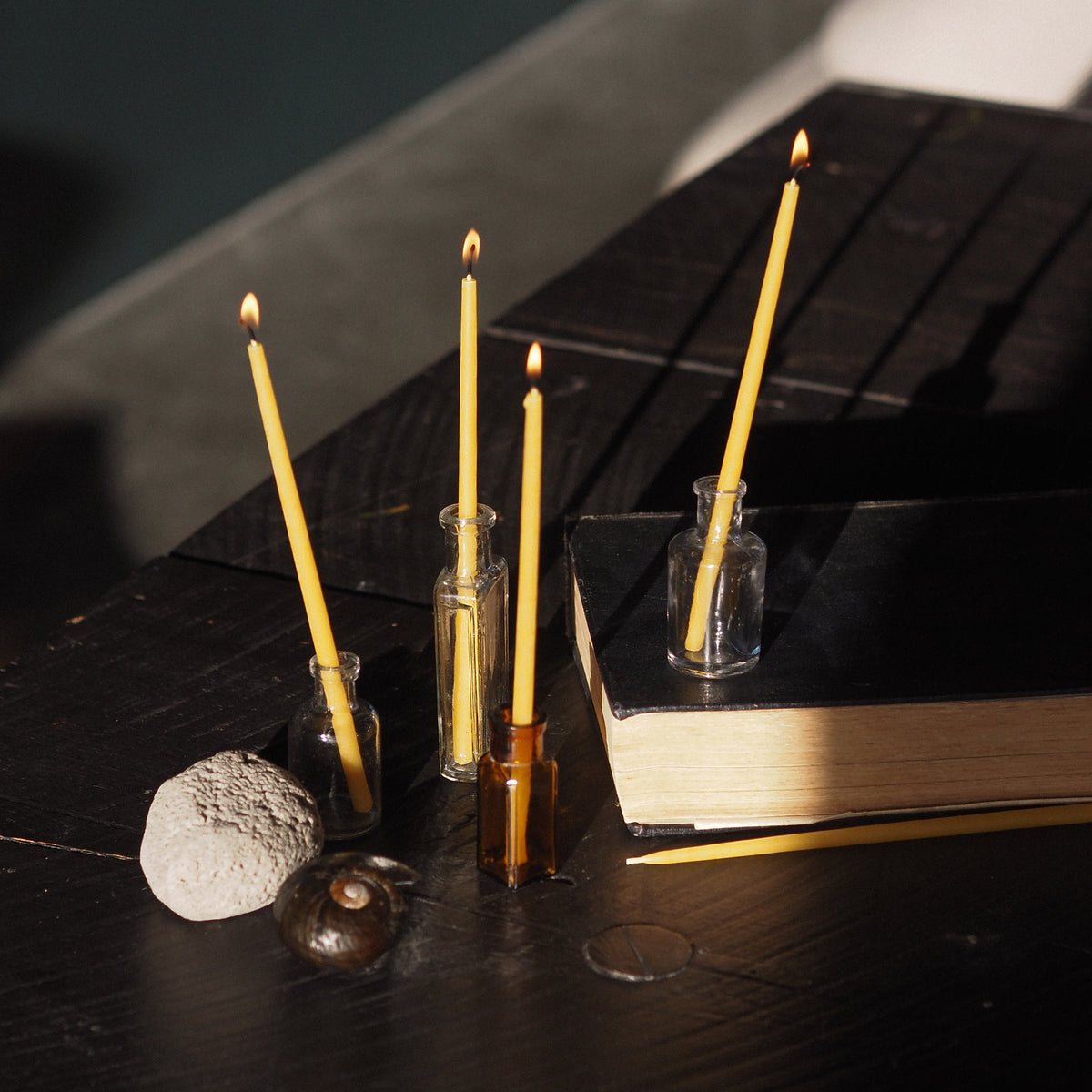 Three lit, thin, beeswax taper candles in small clear glass holders arranged on a dark wooden table beside a black book, with a stone and a small shell nearby in a moody indoor setting.