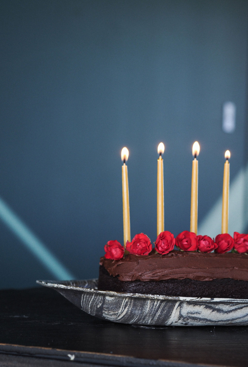 A chocolate cake on a silver marble-patterned plate with five lit candles and red rose decorations, set against a dark blue background.