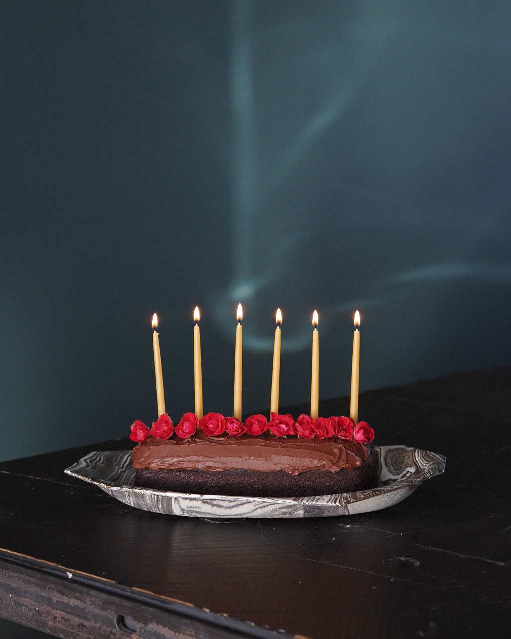 A chocolate cake on a marbled plate with six lit beeswax candles and red rose decorations, set on a dark wooden table against a dark teal background.
