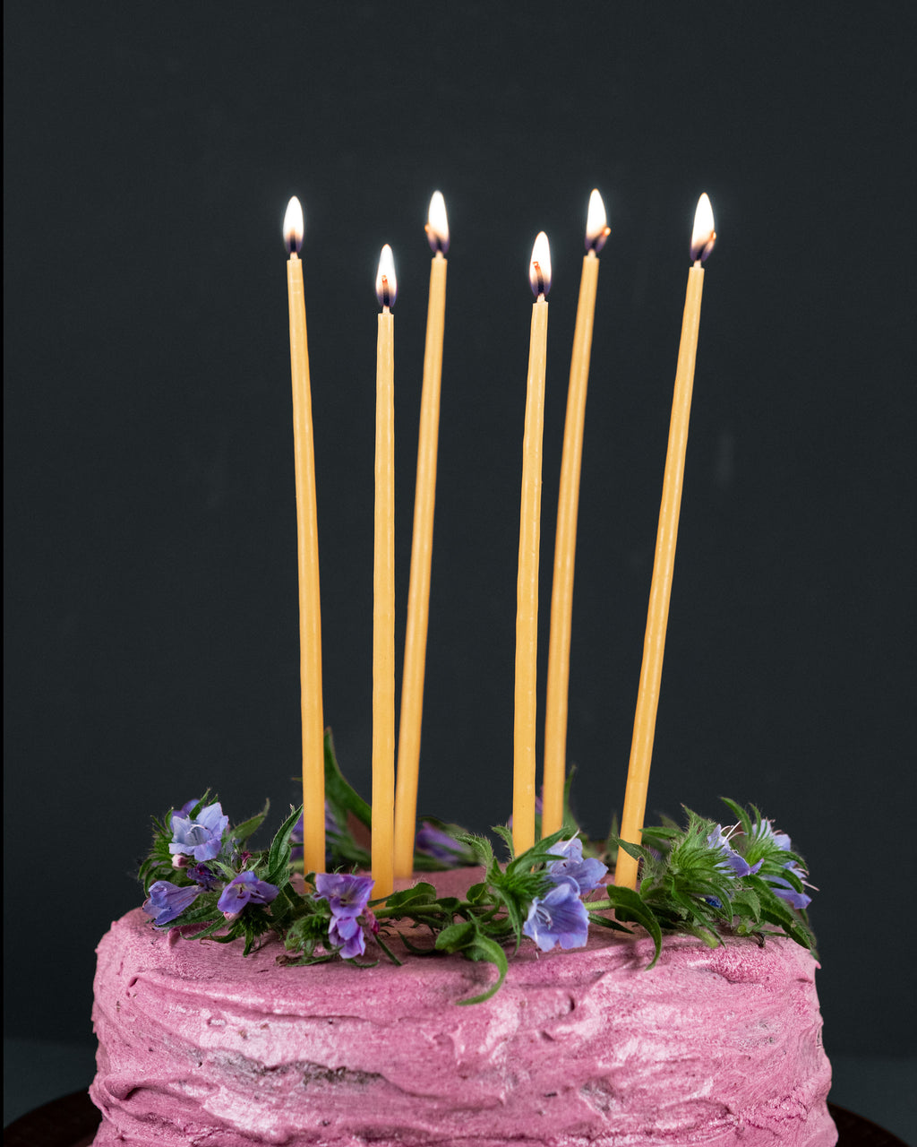 Six tall beeswax birthday candles lit on top of a cake with pink icing and purple flowers. 
