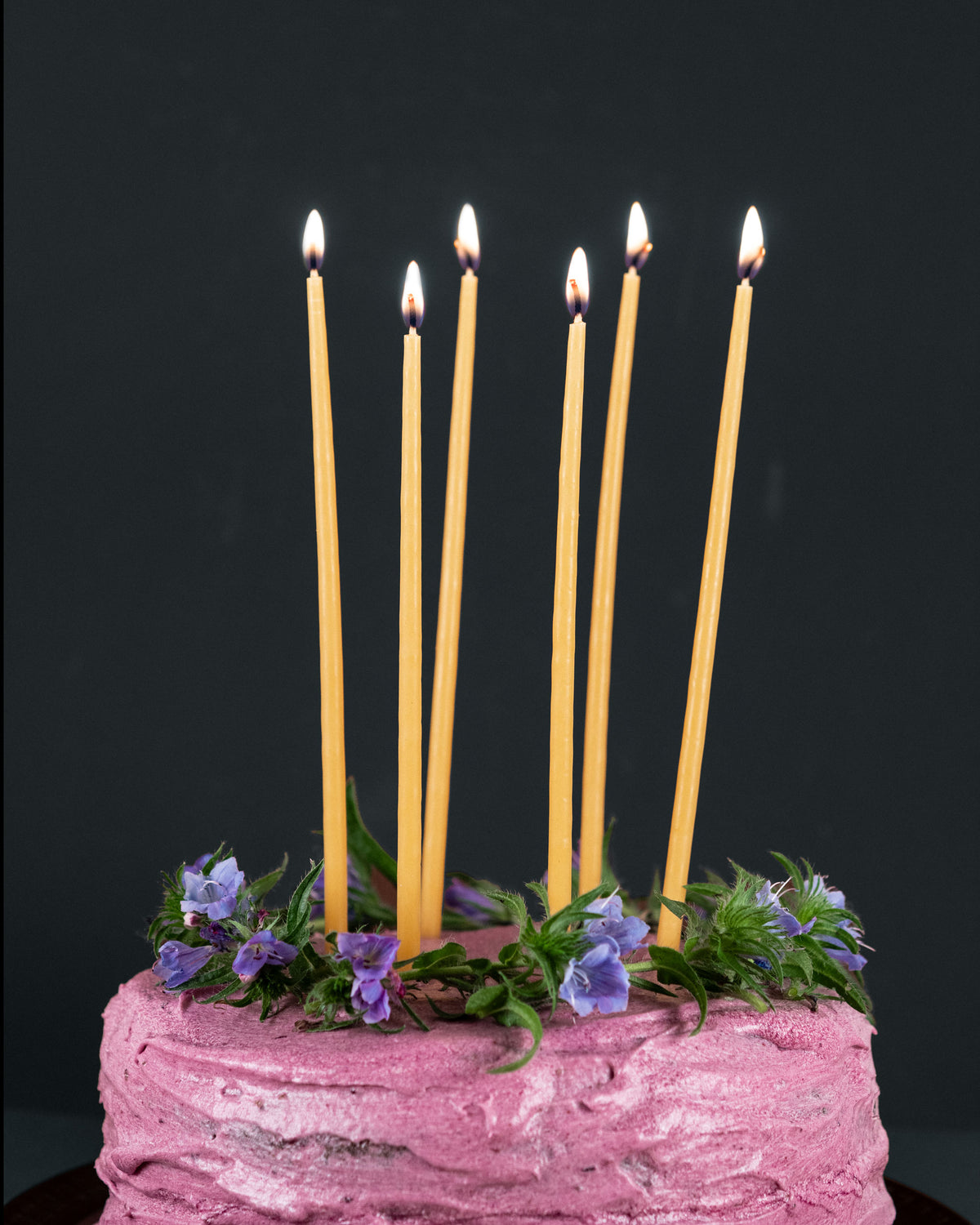 Six tall beeswax birthday candles lit on top of a cake with pink icing and purple flowers. 