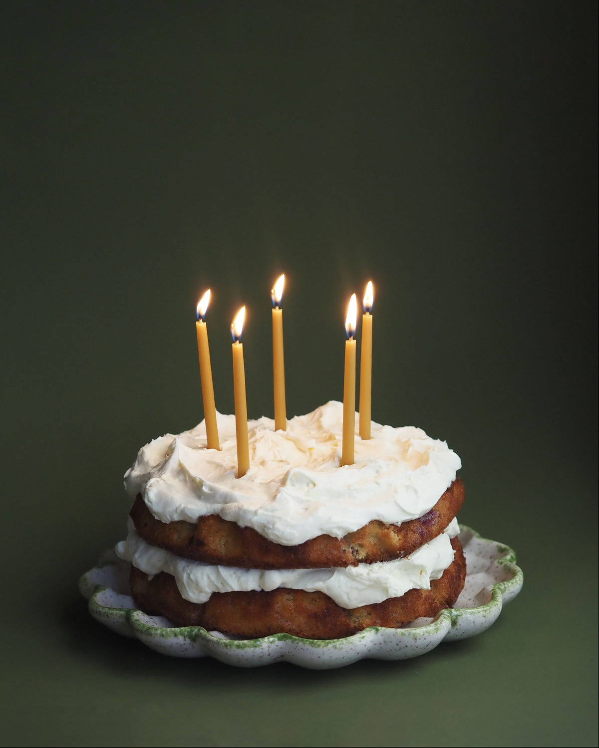 A two-tiered cake with white icing and five lit beeswax candles on top, presented on a scalloped white ceramic plate against a dark olive green background.