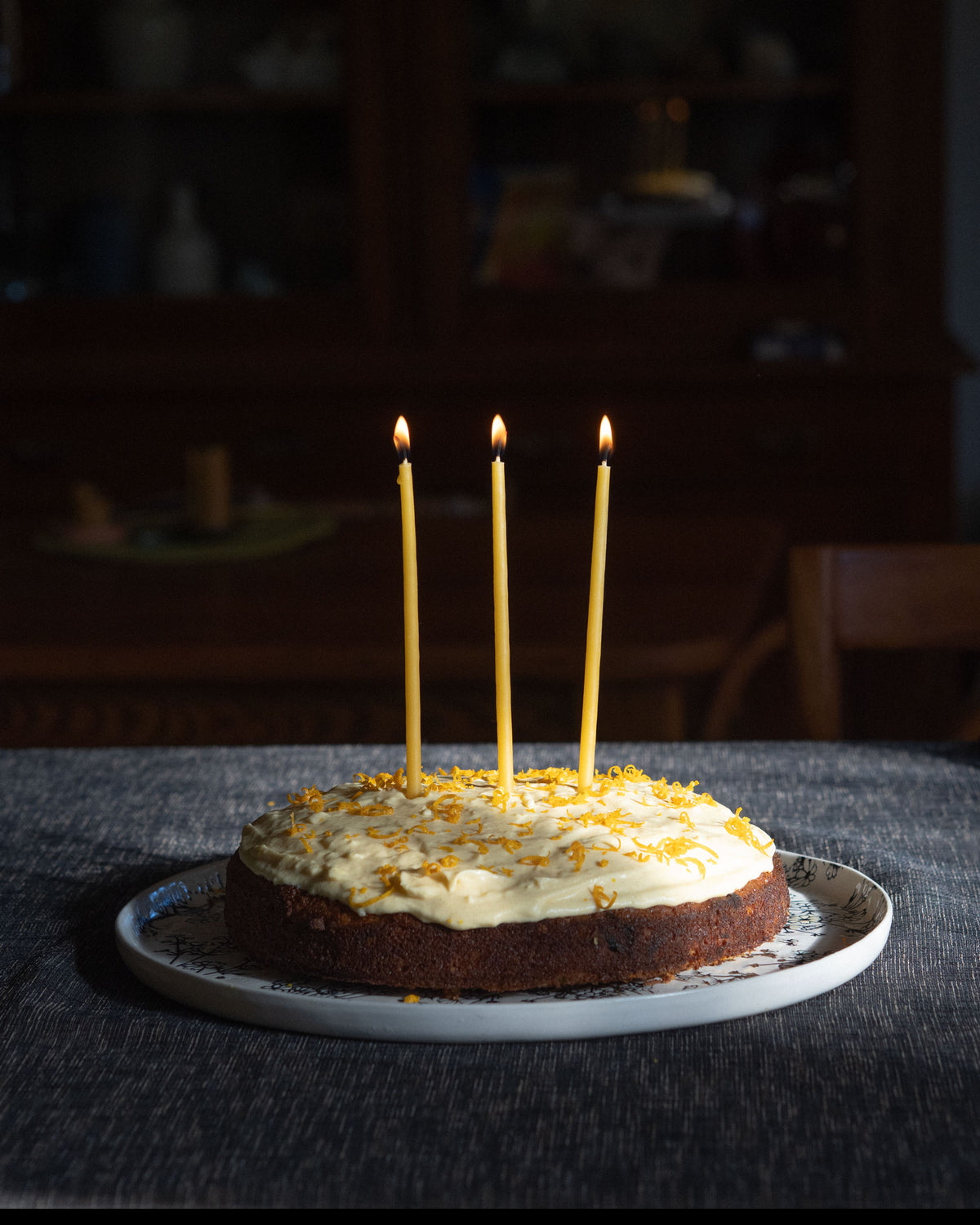 Three lit, thin, beeswax taper candles in a cake with cream icing and orange zest set on a table in a moody indoor setting.