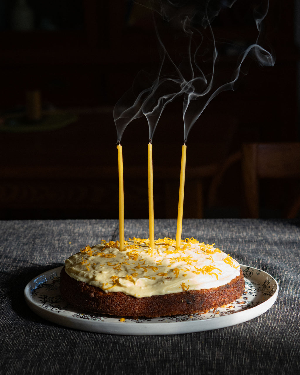 Three thin beeswax birthday candles that have been blown out with smoke rising in a cake with cream icing and orange zest set on a table in a moody indoor setting.