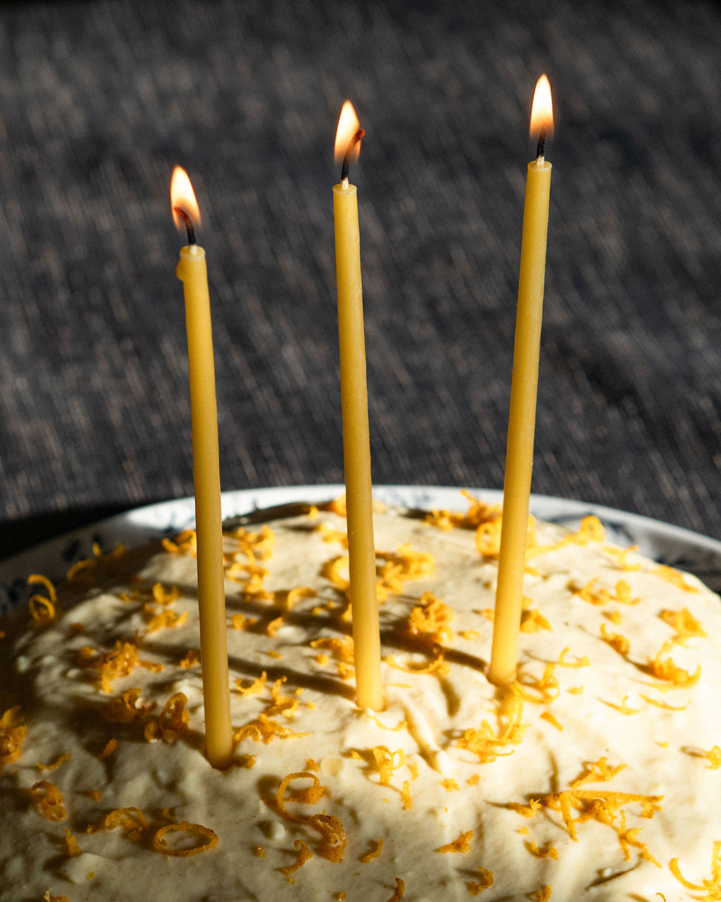 Three lit, thin, beeswax taper candles in a cake with cream icing and orange zest
