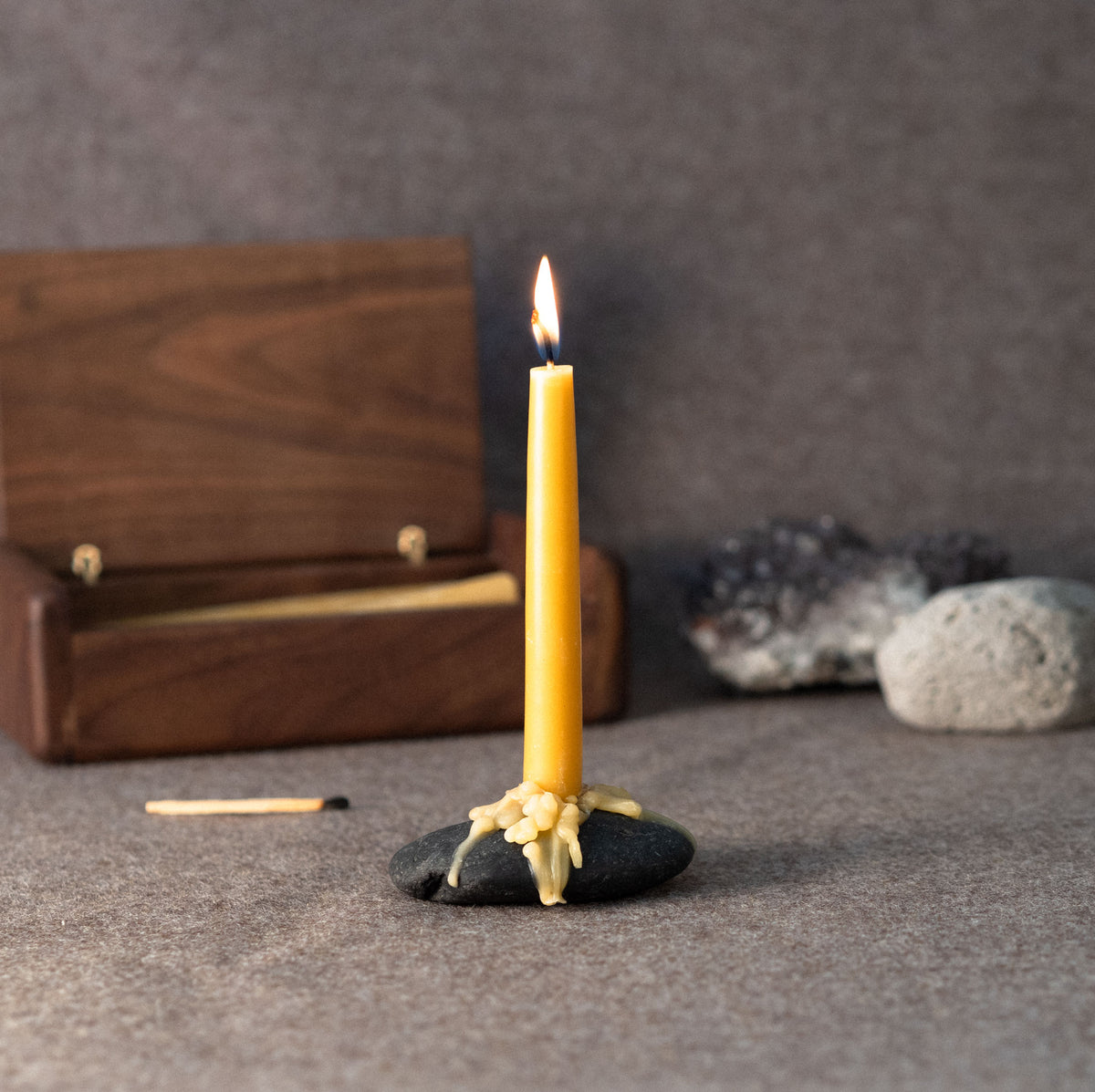 A lit golden beeswax chime candle on a small black stone with wax dripping over it, set on a textured gray surface with a wooden box and stones in the background.