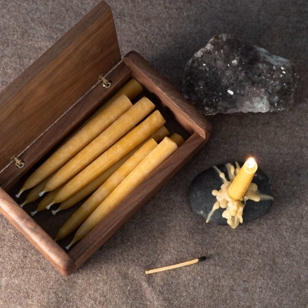 A wooden box filled with small beeswax taper candles, shown on a textured gray surface with a lit chime candle on a small black stone, a crystal cluster, and a match nearby.