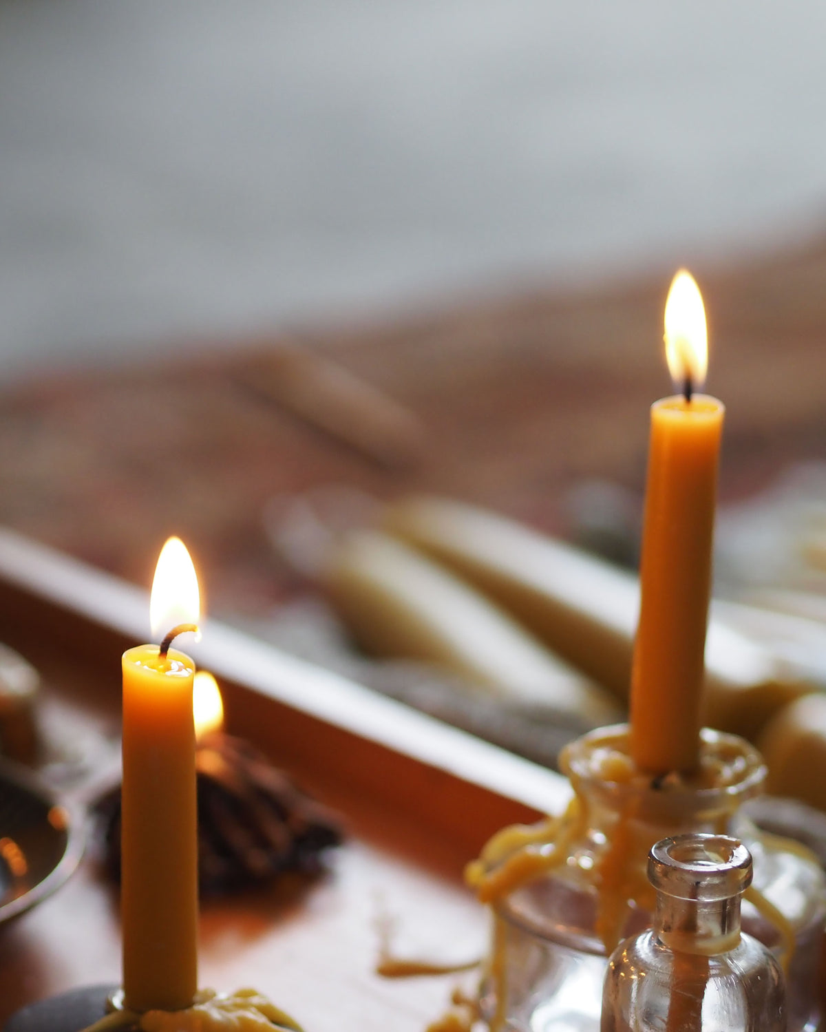 Two lit yellow taper candles in glass holders on a wooden surface, with a softly blurred background showing more candles.