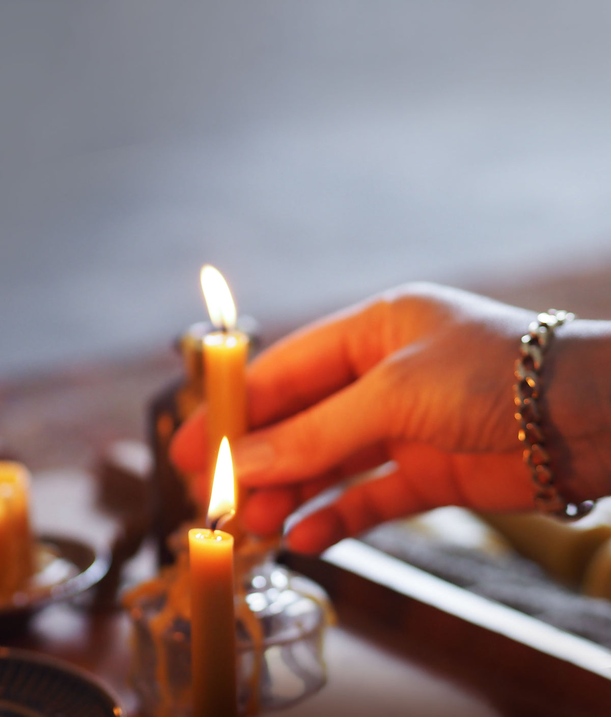 A close-up of a hand lighting a small lit beeswax candle, with warm orange light on the skin and a blurred background.
