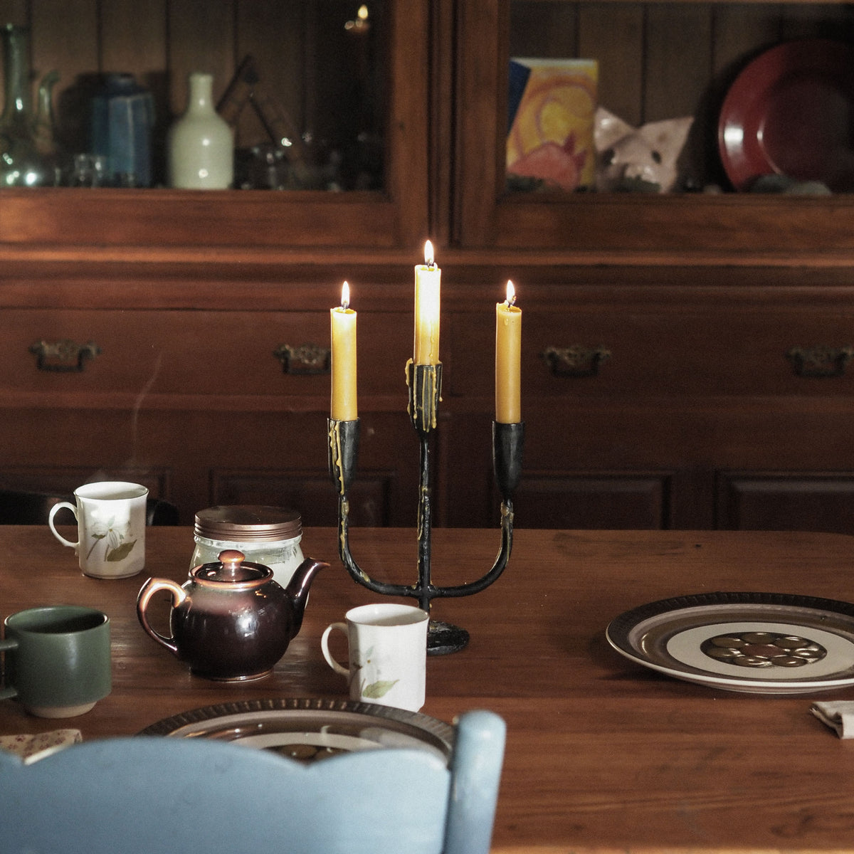A three-arm black metal candelabra holding three lit yellow taper candles sits on a wooden dining table, surrounded by plates, mugs, a teapot, and a blue chair in a warm, rustic room with a wooden hutch in the background.