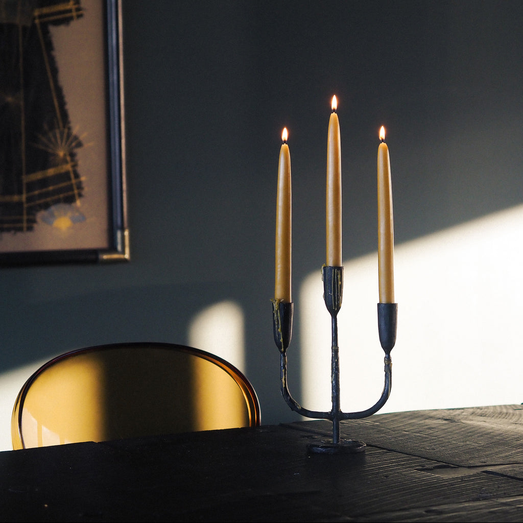 A three-arm metal candelabra holding three lit taper candles sits on a dark wooden table in a dimly lit room, with a chair and framed wall art visible in the background.